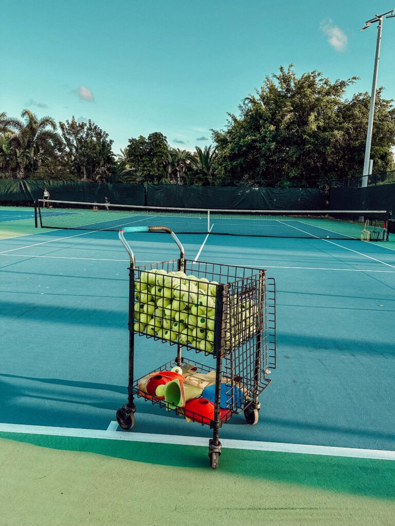 A tennis ball cart stands on a vibrant outdoor tennis court under a clear blue sky, surrounded by greenery.